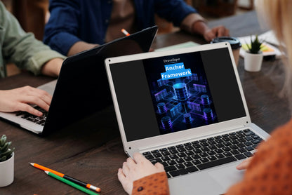 A person at a wooden desk using a silver laptop that displays a presentation slide titled 'Draveliqor Anchor Framework' with blue and purple digital graphics. Other people and laptops are visible in the background of the office setting.