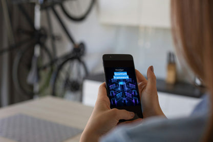 A person holding a black smartphone showing a blue and purple digital interface, with a blurred indoor background and a bicycle visible behind them.